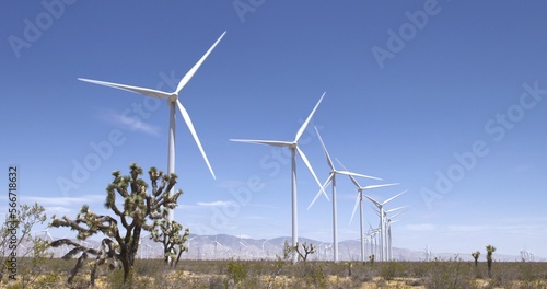 Wide view of turbines operating on scrubby desert land at a wind farm near the Tehachapi Mountains in California.  Blue sky with a few clouds in the distance.