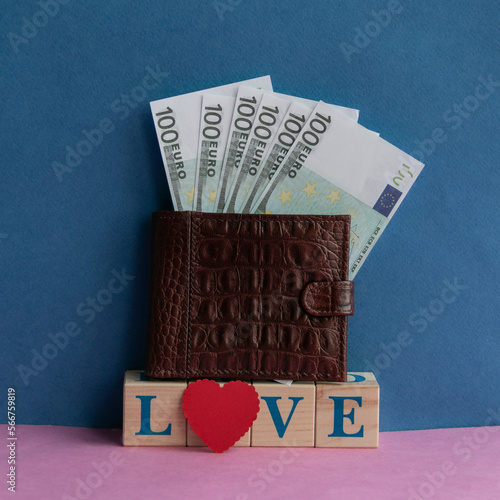 Leather wallet on cubes with the inscription: LOVE and a red heart on a pink-blue background. Euro sticking out of the wallet. Cash donations