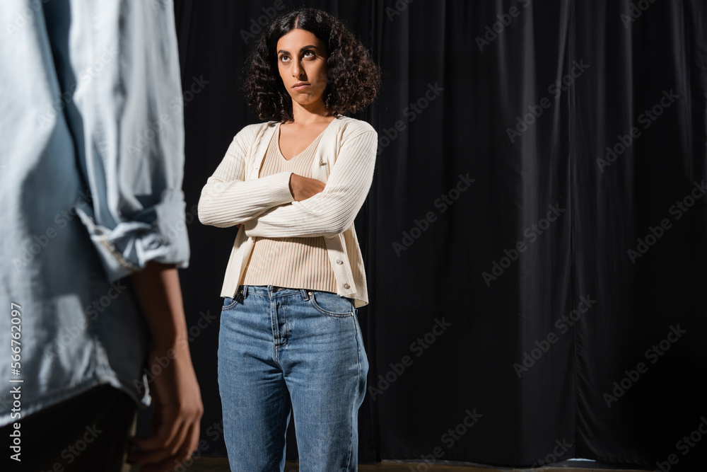 multiracial actress with displeased face expression standing with crossed arms during rehearsal near blurred african american woman. multiracial actress with displeased face expression standing with crossed arms during rehearsal near blurred african american woman.