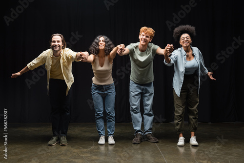 full length of happy multiethnic actors holding hands and bowing on stage of theater.