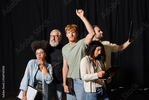 redhead man standing with raised hand near bearded art director and interracial actors with clipboards.