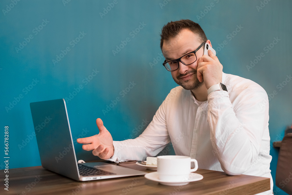 A young European attractive businessman with glasses and a beard speaks on the phone working in a laptop during a coffee shop A young European attractive businessman with glasses and a beard speaks on the phone working in a laptop during a coffee shop