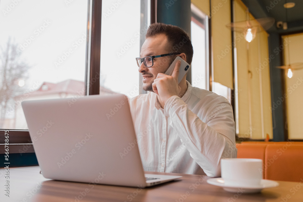 Portrait of a satisfied businessman in a white shirt, glasses, with a beard, talking on the phone and looking at a laptop with a cup of coffee in a cafe Portrait of a satisfied businessman in a white shirt, glasses, with a beard, talking on the phone and looking at a laptop with a cup of coffee in a cafe