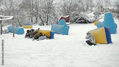 Slow motion video of paintball players in winter who run through snow and hold paintball guns in their hands and shoot from them. General shot of players in camouflage and military uniform in nature