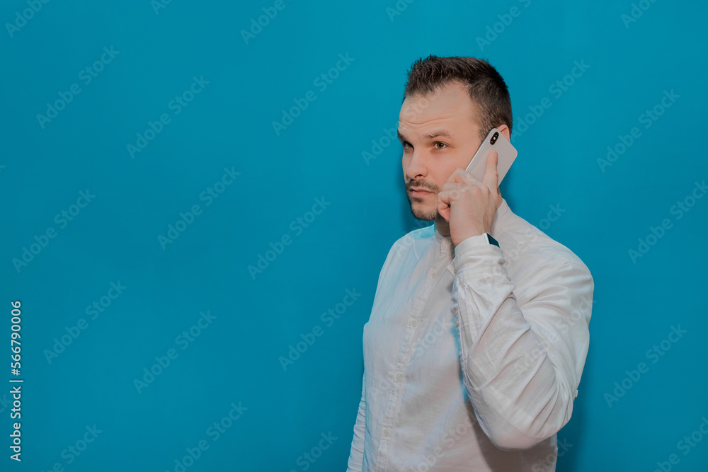 Stylish business serious guy businessman of European appearance in a white shirt speaks on a smartphone against the blue wall background Stylish business serious guy businessman of European appearance in a white shirt speaks on a smartphone against the blue wall background