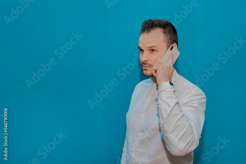 Stylish business serious guy businessman of European appearance in a white shirt speaks on a smartphone against the blue wall background