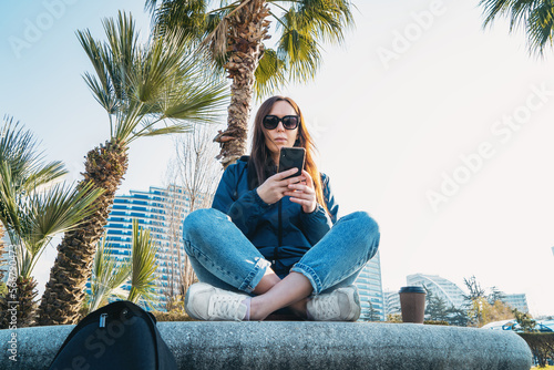 Young beautiful brunette girl in sunglasses use smartphone while sitting cross-legged in city park.