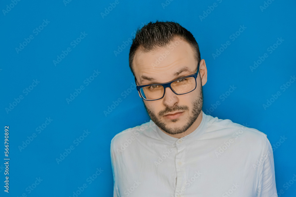 Portrait of a stylish young guy Caucasian businessman with frown eyebrows, glasses and a white shirt on a blue background, close-up Portrait of a stylish young guy Caucasian businessman with frown eyebrows, glasses and a white shirt on a blue background, close-up