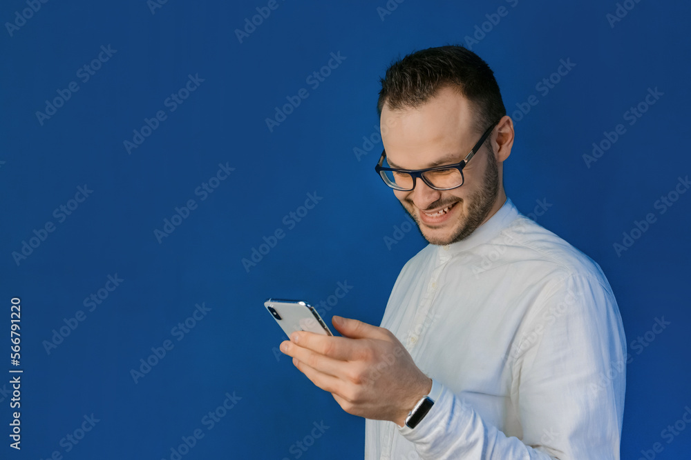 Portrait of a stylish, smiling businessman from Europe in glasses and a white shirt, spending time in his mobile phone on a blue wall background Portrait of a stylish, smiling businessman from Europe in glasses and a white shirt, spending time in his mobile phone on a blue wall background