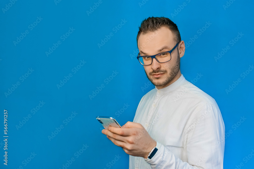 Stylish, businesslike, confident young guy businessman from Europe, wearing glasses and white shirt, holds a smartphone on a blue background of the wall Stylish, businesslike, confident young guy businessman from Europe, wearing glasses and white shirt, holds a smartphone on a blue background of the wall