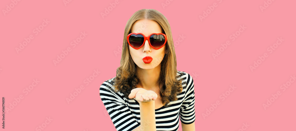 Portrait of beautiful young woman blowing her lips with lipstick sending sweet air kiss wearing red heart shaped sunglasses on pink background Portrait of beautiful young woman blowing her lips with lipstick sending sweet air kiss wearing red heart shaped sunglasses on pink background