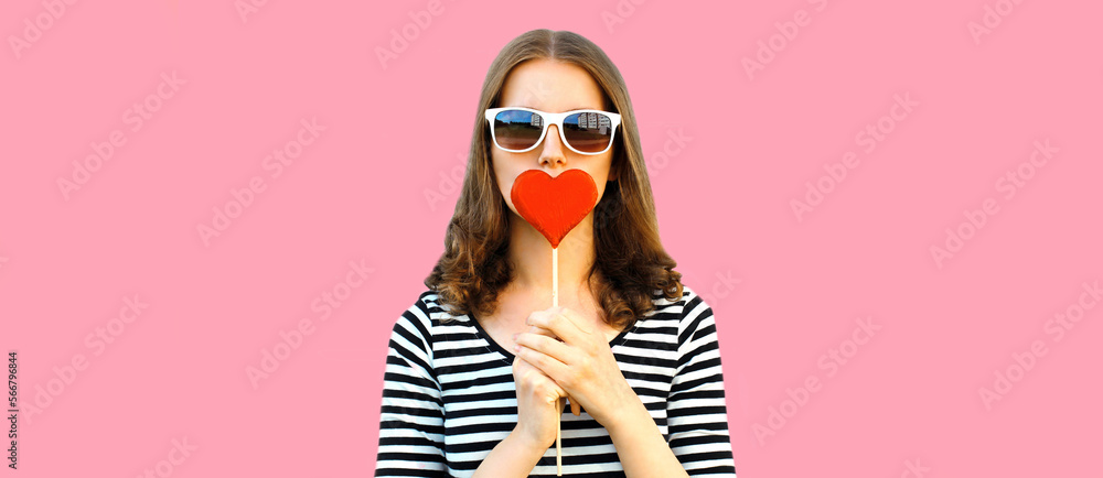 Portrait of beautiful young woman covering her lips with red sweet heart shaped lollipop on pink background Portrait of beautiful young woman covering her lips with red sweet heart shaped lollipop on pink background