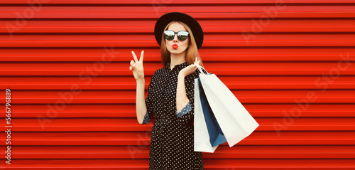 Portrait of beautiful young woman with shopping bags blowing her lips sends air kiss wearing black round hat on red background © rohappy