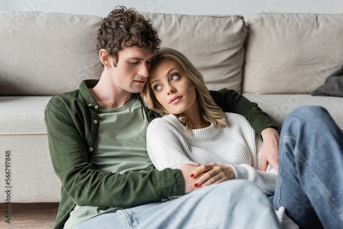 young man with curly hair hugging blonde woman near sofa in living room.