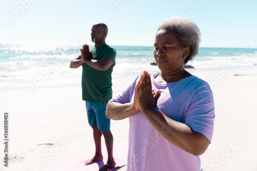 African american senior man and woman meditating in prayer pose at beach against clear sky