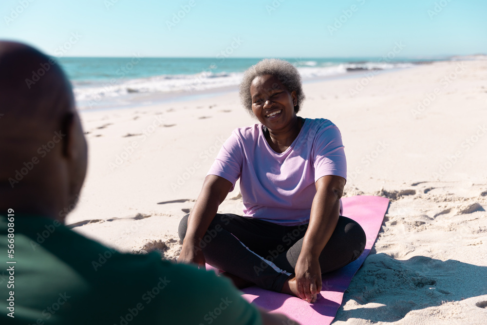 Happy african american senior woman talking with man while relaxing on mats at beach in summer