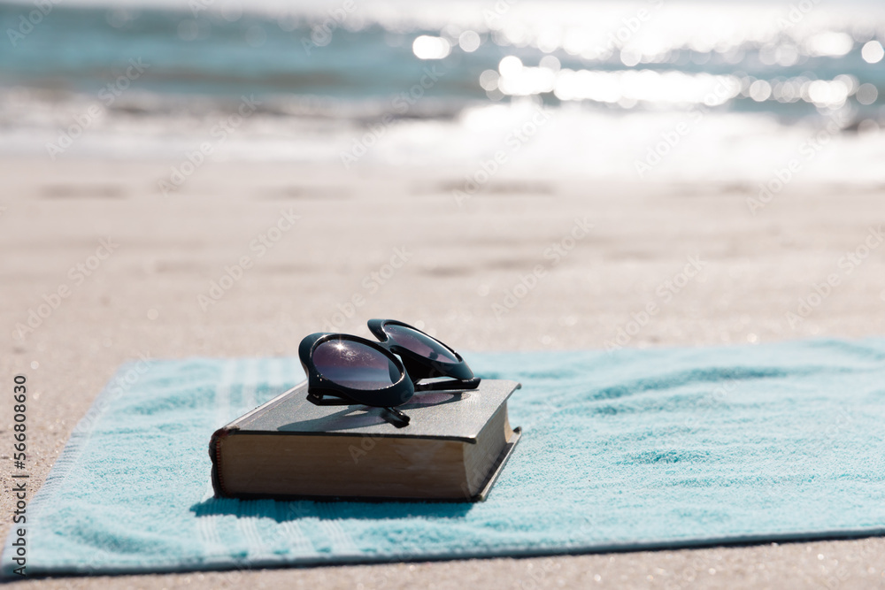 Obraz premium Close-up of book with sunglasses and blue towel on sandy beach against sea during sunny day