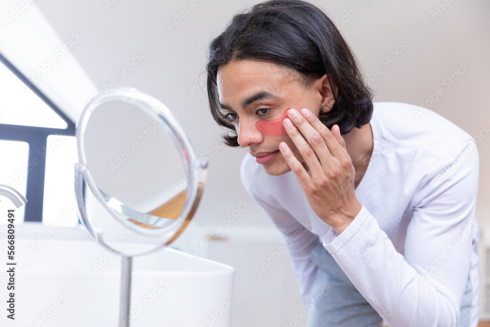Happy biracial man looking in mirror, applying eye patches in bathroom Happy biracial man looking in mirror, applying eye patches in bathroom