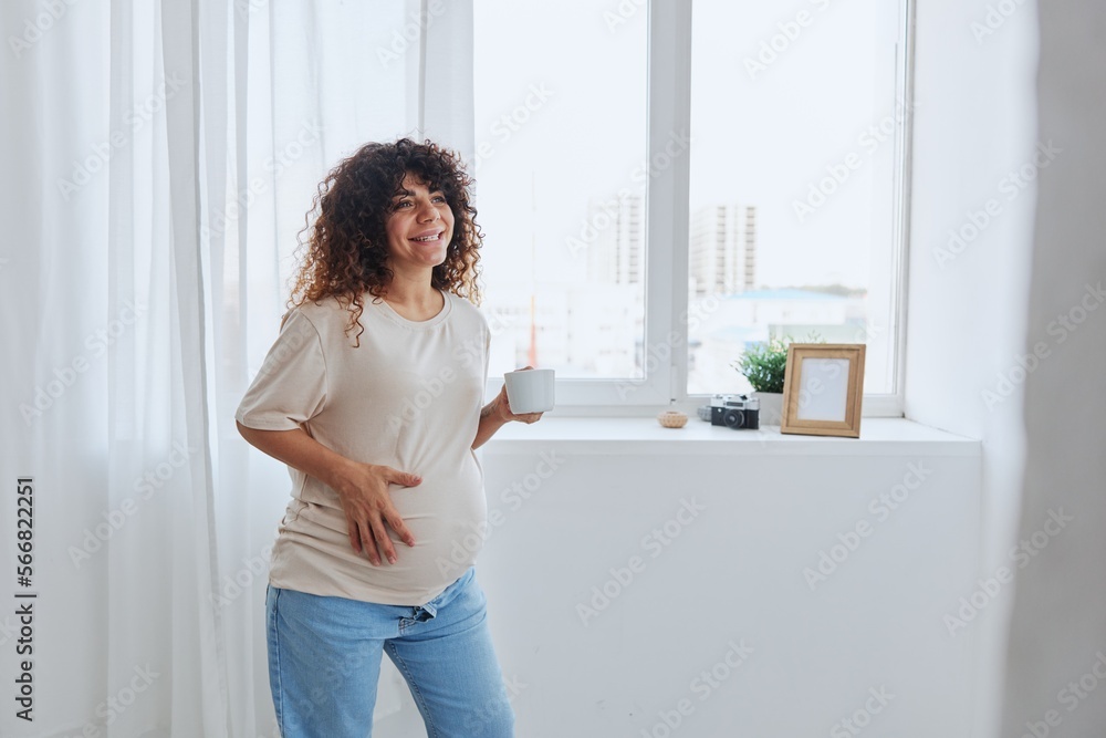 A pregnant woman with curly hair stands at the window with a mug of warm water and tea and enjoys the view in a home t-shirt, the happiness of motherhood A pregnant woman with curly hair stands at the window with a mug of warm water and tea and enjoys the view in a home t-shirt, the happiness of motherhood