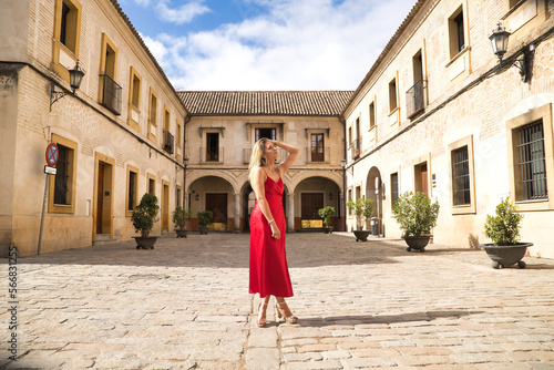 Young, attractive, blonde woman in an elegant red party dress sensually touching her hair in the city. Concept beauty, fashion, elegance, luxury, hairstyle, sensuality.