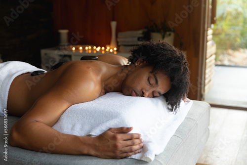 Relaxed biracial man on massage table having hot stones treatment at spa, copy space