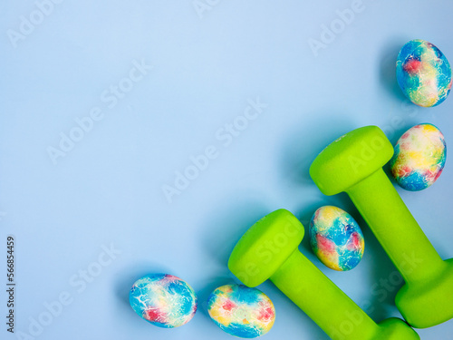 green heavy dumbbells, colorful easter eggs on blue background. Easter fitness and training frame composition with copy space. Top view from above, flat lay.