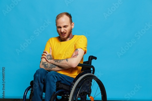 A man in a wheelchair in a T-shirt with tattoos on his arms sits on a blue studio background, a full life, a real person