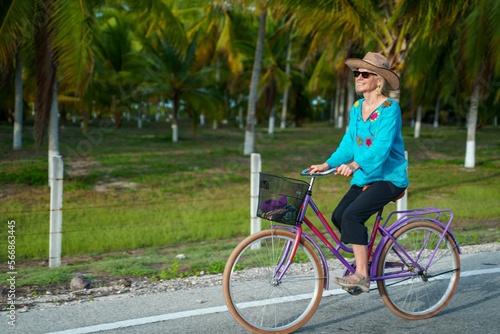 Side view of happy smiling pretty mature senior woman smiling biking towards the camera wearing ethnic clothes and hat and sunglasses on a tropical road.