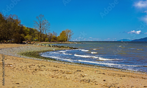 Fototapeta Naklejka Na Ścianę i Meble -  A fragment of a beach with a forest area in the English bay in spring, blue water with small waves against the backdrop of a mountain range and blue sky