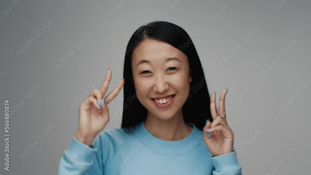 Portrait of Young Korean Woman Looking at Camera in Colour Studio Shot. Adult Japanese Girl Isolated Alone on Grey Background Closeup. Happy 20s Asian Person Smiling and Showing Peace Sign by Fingers Portrait of Young Korean Woman Looking at Camera in Colour Studio Shot. Adult Japanese Girl Isolated Alone on Grey Background Closeup. Happy 20s Asian Person Smiling and Showing Peace Sign by Fingers