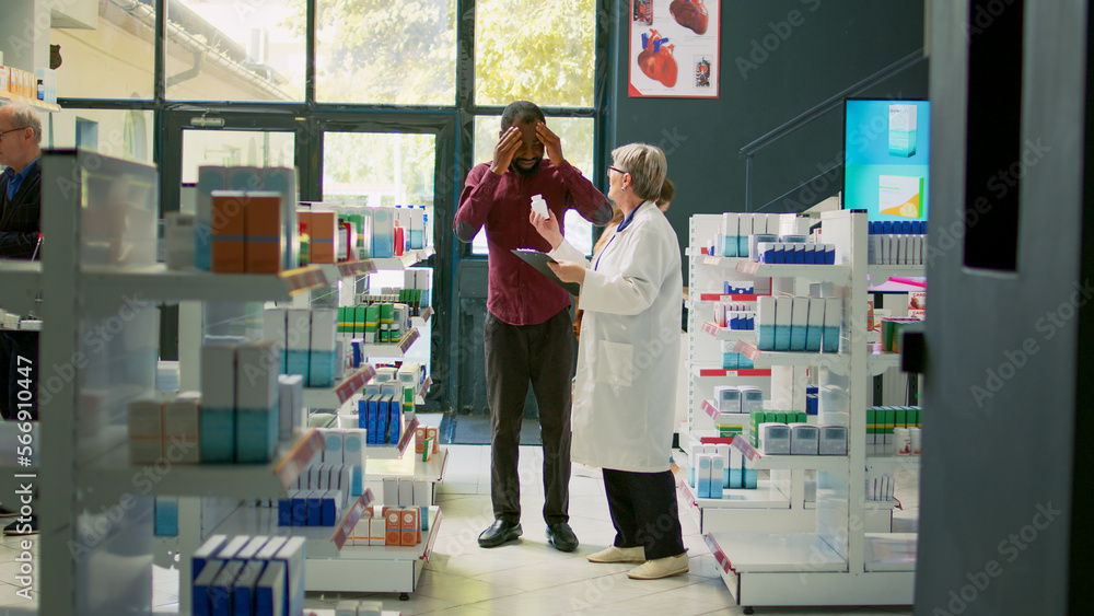 Obraz premium Sick male client entering drugstore with painful headache, asking senior pharmacist about cure or medicaments treatment. Customer suffering from migraine and rubbing temples in drugstore.