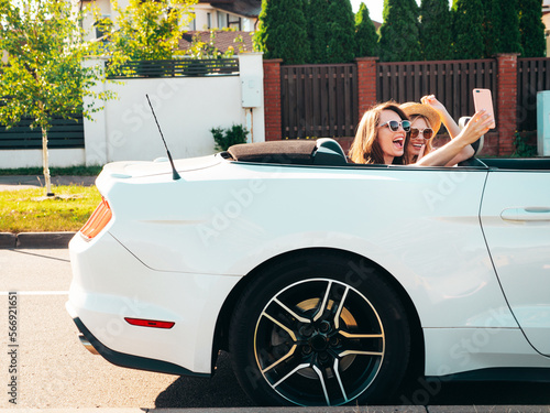 Two young beautiful and smiling hipster female in convertible car. Sexy carefree women driving cabriolet. Positive models riding, having fun in sunglasses outdoors. Enjoying summer days. Take selfie © halayalex