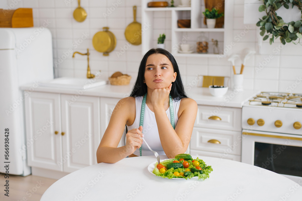 Woman with a measuring tape around her neck is sitting at a table with a dissatisfied face in front of a plate of vegetables. The concept of healthy eating