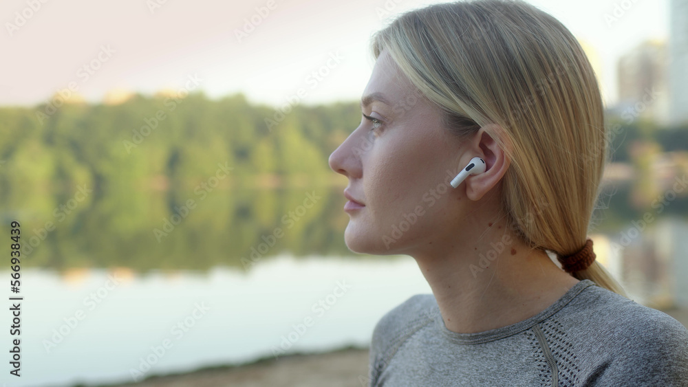 athletic blonde young woman sits on bank of river or lake and puts on headphones to go in for sports while listening to music. Girl getting ready for outdoor sports