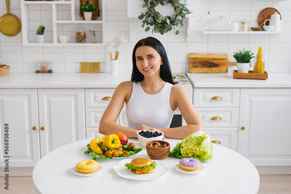 Happy healthy woman is sitting at a table with healthy food, fresh fruits and vegetables in the kitchen Happy healthy woman is sitting at a table with healthy food, fresh fruits and vegetables in the kitchen