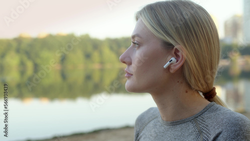 athletic blonde young woman sits on bank of river or lake and puts on headphones to go in for sports while listening to music. Girl getting ready for outdoor sports