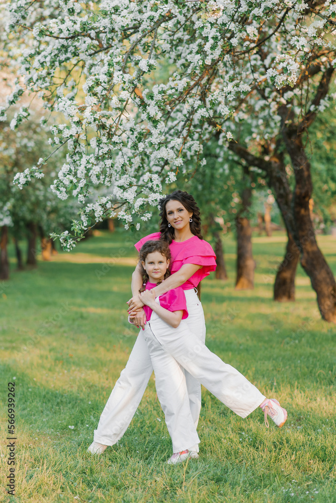 A sweet young woman in white trousers and a bright pink blouse with her daughter is walking through a spring park or garden among the flowering trees of an apple tree and hugs her