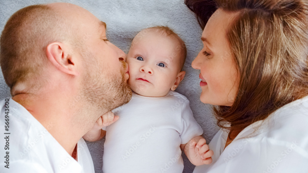 Happy family. Newborn baby with happy parents, top view. Healthy newborn baby in a white t-shirt with mom and dad. Close up Faces of the mother, father and infant baby. Cute Infant boy and parents Happy family. Newborn baby with happy parents, top view. Healthy newborn baby in a white t-shirt with mom and dad. Close up Faces of the mother, father and infant baby. Cute Infant boy and parents
