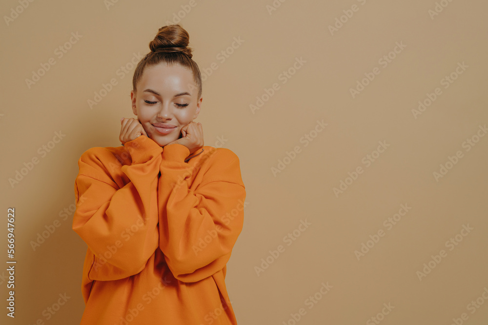 Delightful woman in orange sweatshirt smiling with closed eyes, keeping hands near face and dreaming Delightful woman in orange sweatshirt smiling with closed eyes, keeping hands near face and dreaming