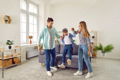 Happy smiling family having fun with their child son in the living room at home. Couple lifting in the air their laughing cheerful boy kid in pilot's glasses and playing plane with him.