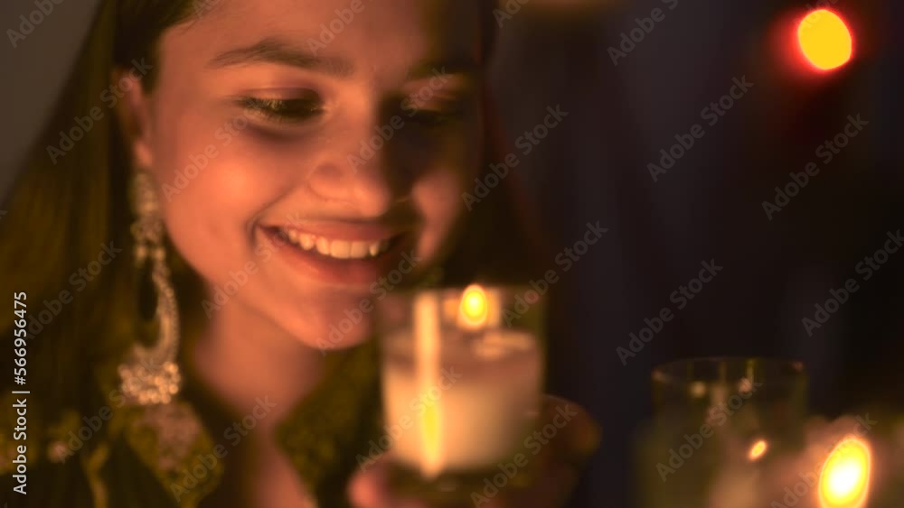 Smiling pretty Indian teen sitting and holding burning glass candle in hand on the occasion of Diwali festival. Girl enjoying Deepavali with Decorative surroundings , burning Diya at home.