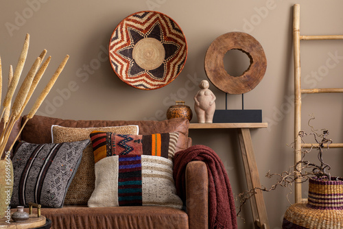 Canvas Print Interior design of ethnic living room interior with colorful pillows, brown sofa, wooden bench, sculpture, vase with dried flowers, ladder, claret plaid and personal accessories