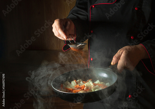 Fototapeta Naklejka Na Ścianę i Meble -  The chef adds paprika to fried carrots and onions in a frying pan. Cooking vegetables for lunch. Restaurant or hotel menu on a dark background