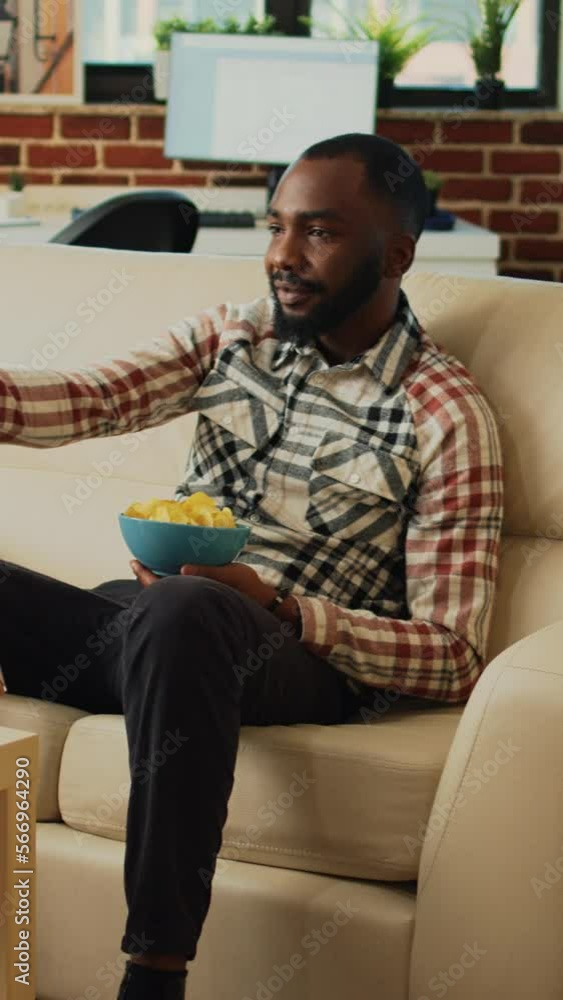 Vertical video: Male adult eating chips from bowl and drinking beer, laughing at comedy film while he has fast food meal on table. Young man serving noodles, hamburger and pizza in living room. Vertical video: Male adult eating chips from bowl and drinking beer, laughing at comedy film while he has fast food meal on table. Young man serving noodles, hamburger and pizza in living room.