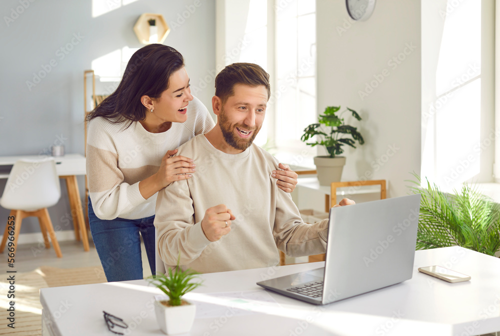 Family couple happy about a good job offer. Young man and woman looking at their laptop computer with excited face expressions. Wife hugs her husband by the shoulders when they learn about his success Family couple happy about a good job offer. Young man and woman looking at their laptop computer with excited face expressions. Wife hugs her husband by the shoulders when they learn about his success