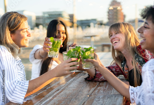 Multiethnic friends raising their mojitos in celebration while enjoying a day at the beach. The girls are sitting at chiringuito bar with the sea in the background. photo