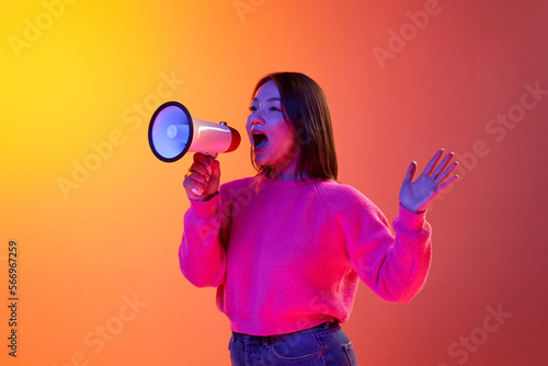 News, information. Young girl in pink sweater talking in megaphone over gradient orange background in neon light. Concept of emotions, facial expression, youth, lifestyle, inspiration, sales, ad