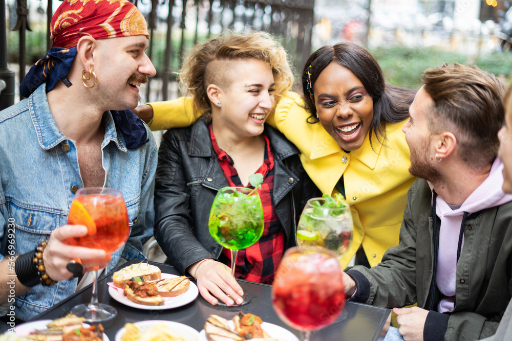  Group of multiethnic friends toasting with bright and colorful cocktails. Friends sitting at a bar table, laughing and having a great time together