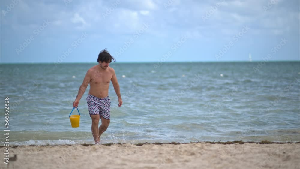 Slow motion of a hispanic man gathering water in the sea for his son walking on the beach wearing short Slow motion of a hispanic man gathering water in the sea for his son walking on the beach wearing short