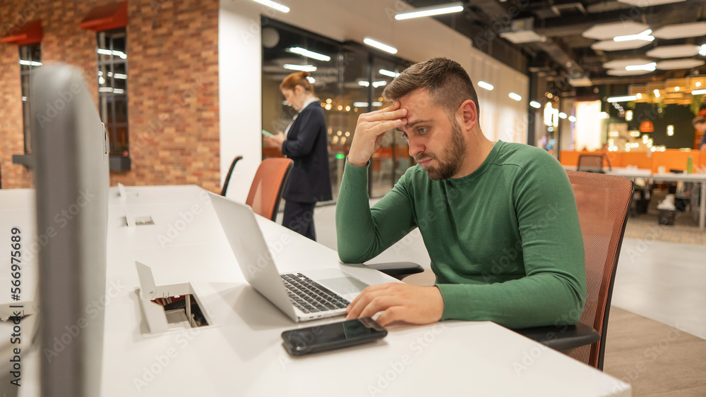 Bearded man holding his head while working on a laptop in an open space office. Red-haired caucasian woman uses smartphone. Bearded man holding his head while working on a laptop in an open space office. Red-haired caucasian woman uses smartphone.